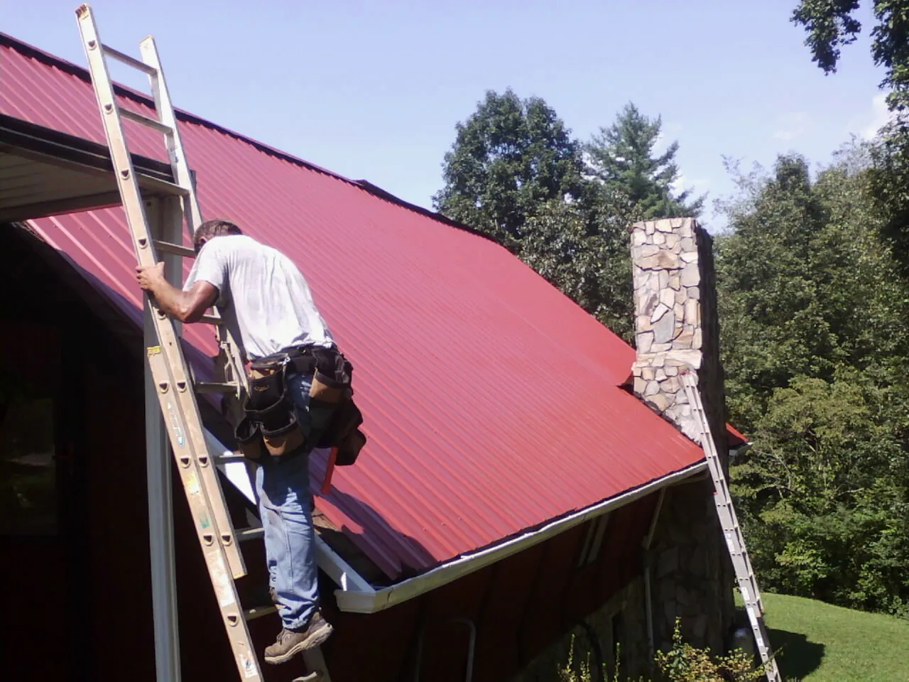 Roofer on a ladder beside a red metal roof and stone chimney — Soffit & Fascia Repair by Roofing Brothers in Jamesville